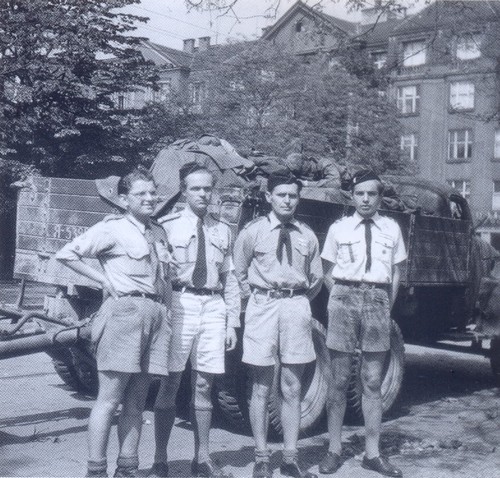 Fotografie čtyř skautů, v pozadí vojenská technika